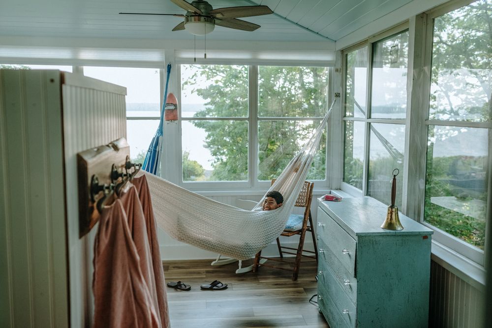Happy boy lays in hammock on a white cabin glass windowed porch. Southern Maine Acadia National Park family vacation photographer.