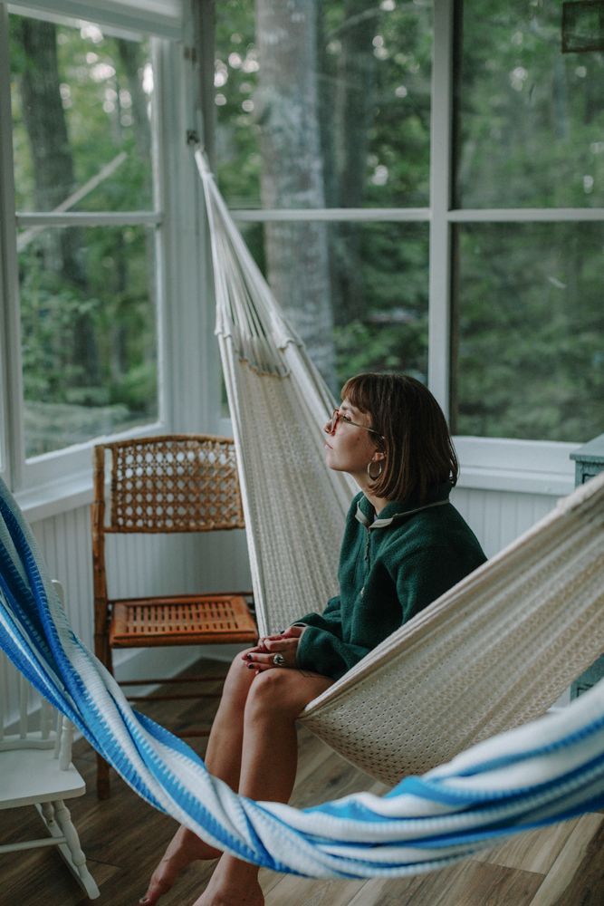 Teen girl sits pensively in a hammock looking out window on a white cabin porch in Acadia National Park. Acadia family vacation photographer