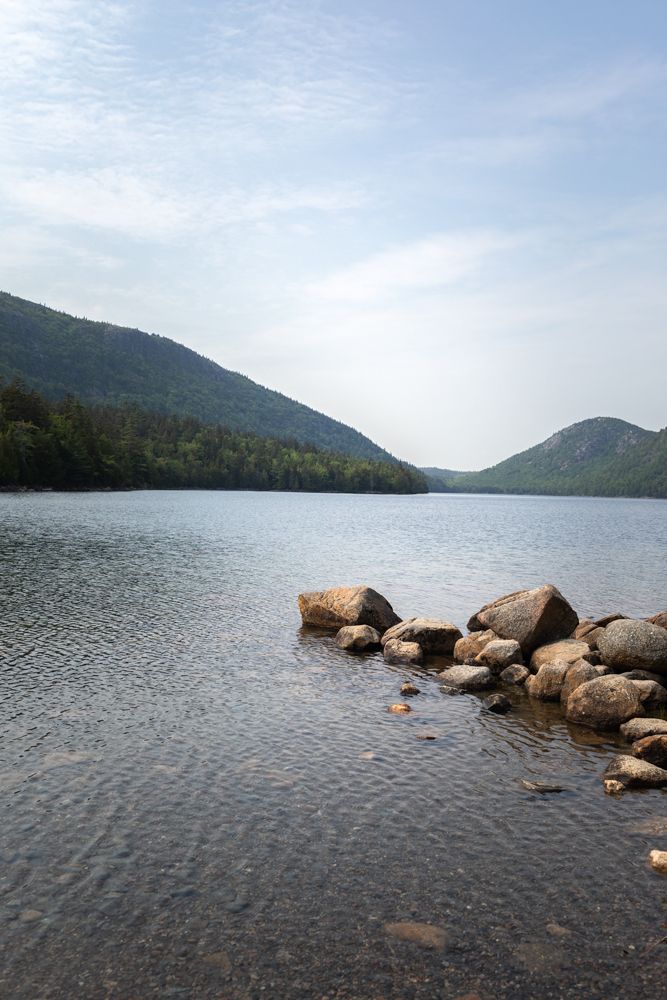 Tranquil mountain lake with rocky shore, pines, and cloudy sky at Jordan Pond. Acadia National Park family vacation photographer.