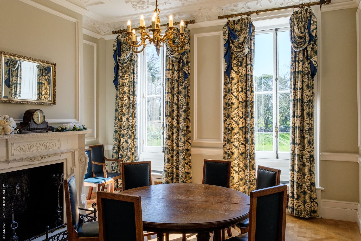 Elegant dining room with ornate blue and gold curtains, round wooden table, fireplace, and chandelier in a classic interior.