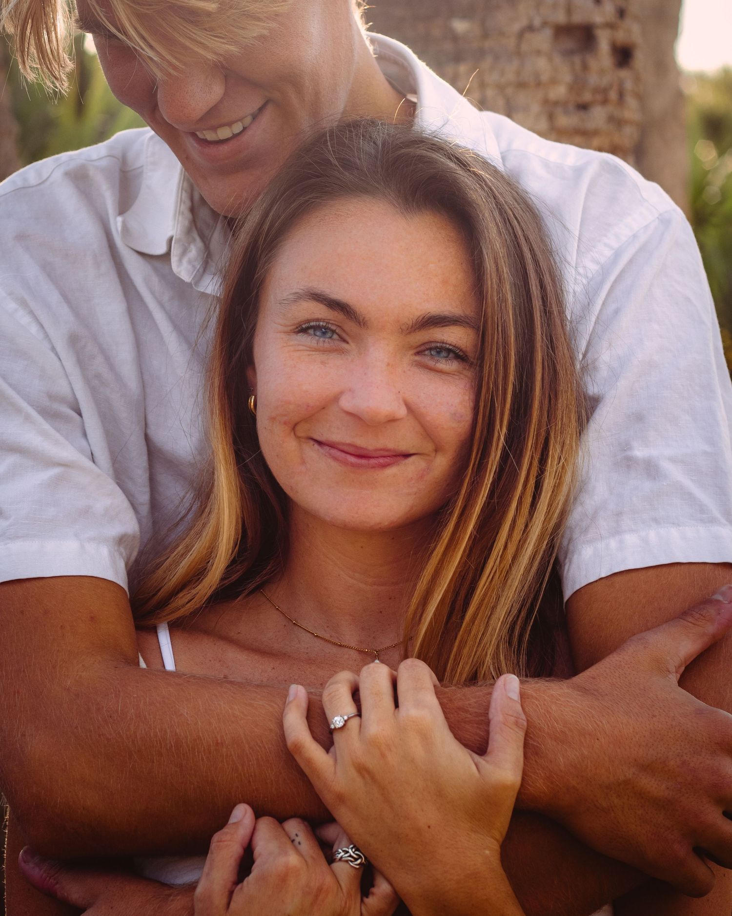 A couple embraces outdoors, woman with highlighted hair and blue eyes smiles warmly while man wraps his arms around her.