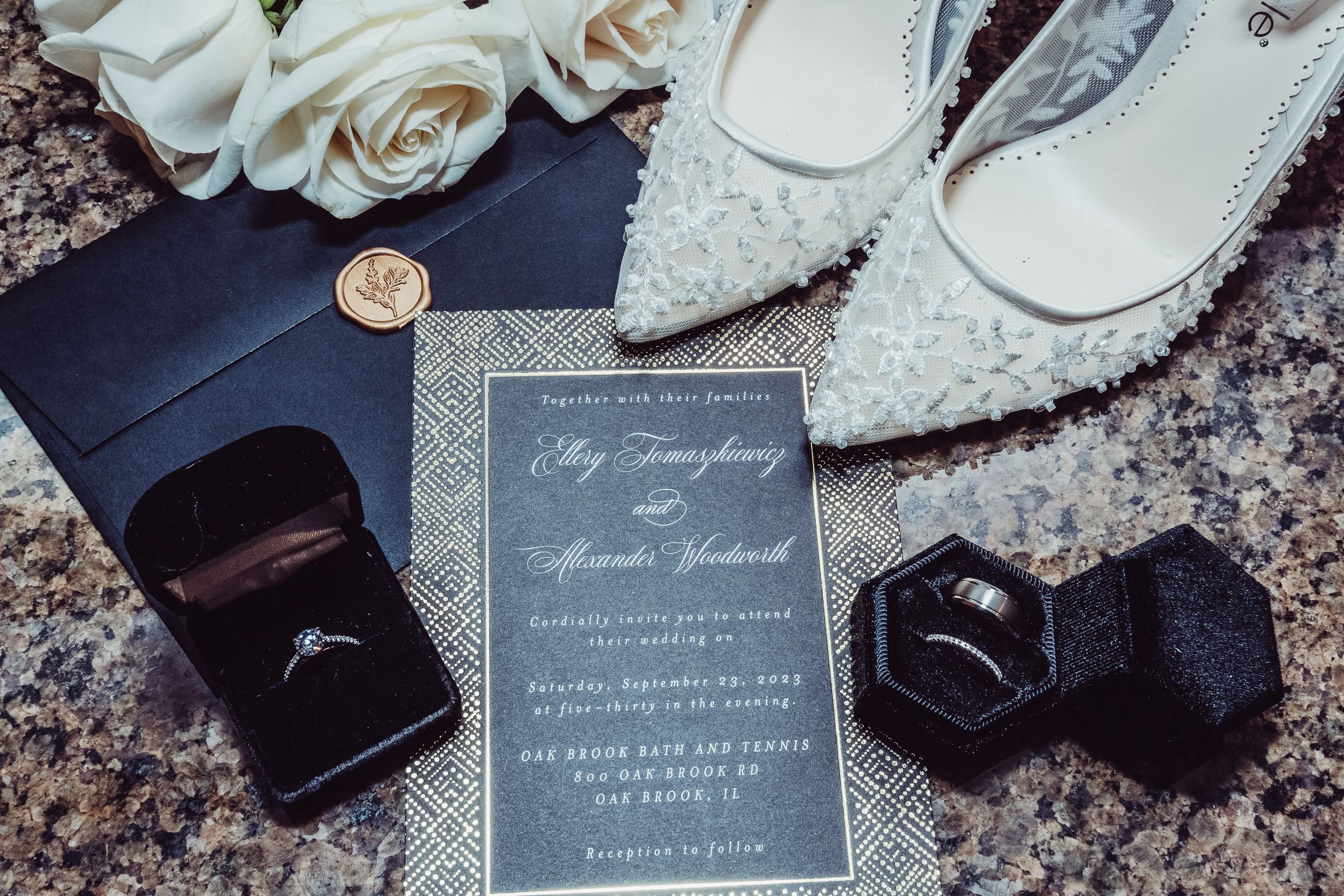 Elegant wedding flat lay with navy invitation, diamond ring, wedding band, white heels, and roses on marble surface.