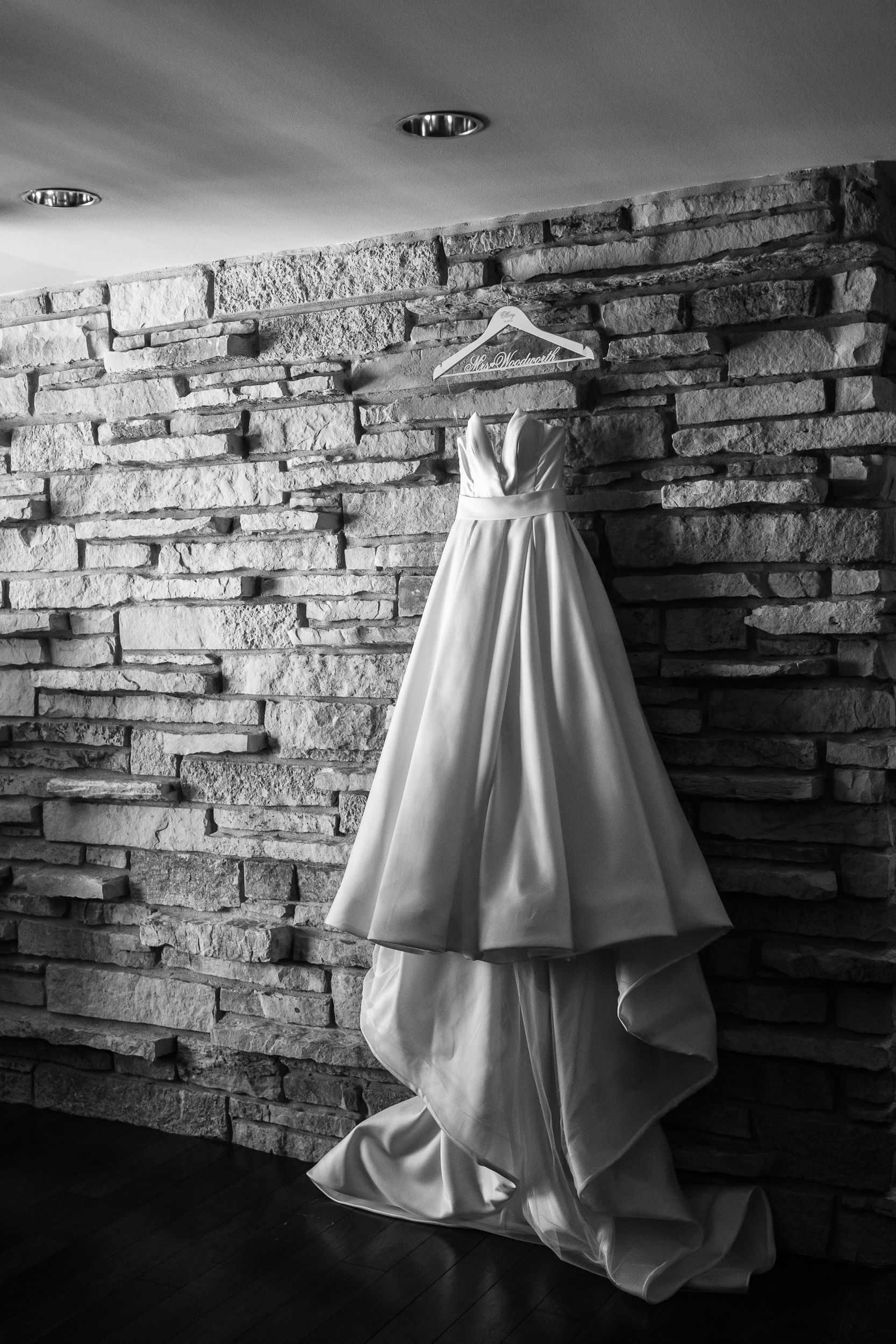 Black and white photo of an elegant wedding gown hanging on a wooden hanger against a rustic stone wall.