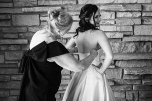 Black and white photo of a woman helping a bride fasten her wedding gown near a stone wall.