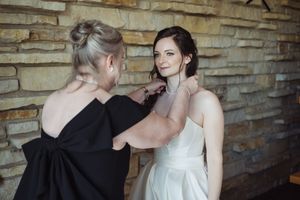 A woman in black dress helps bride in white gown fasten necklace against stone wall backdrop.