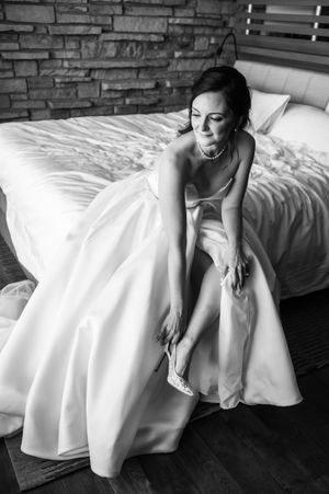 Bride in a white ballgown sitting on a bed putting on her wedding shoes, black and white photo.