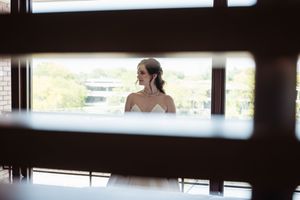 Bride in white dress gazing out large windows, framed by dark wooden slats in a beautifully lit venue.