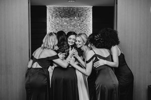 Bridesmaids share an emotional group hug with the bride in a black and white wedding photo.