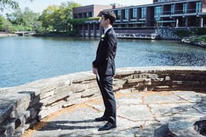 Groom in black tuxedo stands by a stone wall overlooking a river in an outdoor wedding portrait.