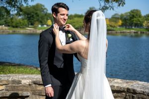 Bride in white gown adjusting groom's boutonniere by a scenic lakeside on a sunny wedding day.