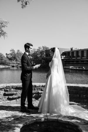 Groom reads vows to bride in wedding dress and veil by a scenic waterfront in black and white.