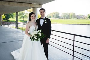 Bride and groom pose together on waterfront promenade, bride holding white floral bouquet with greenery.