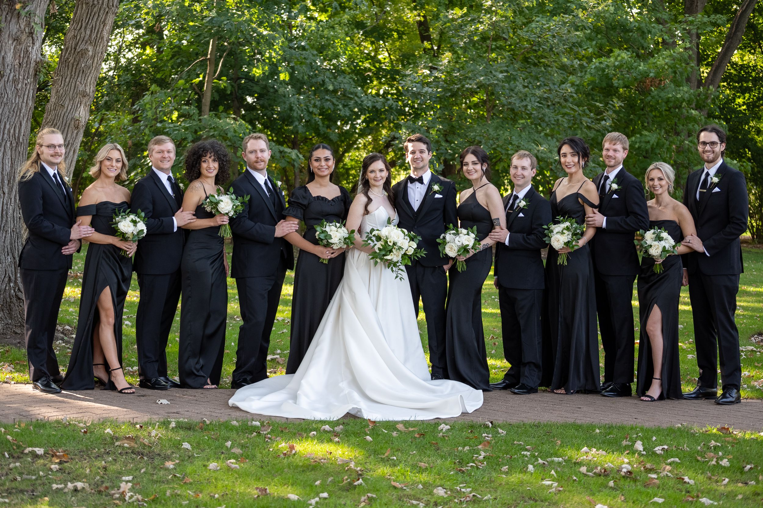 Wedding party poses in matching black attire with bride and groom in a lush outdoor setting.