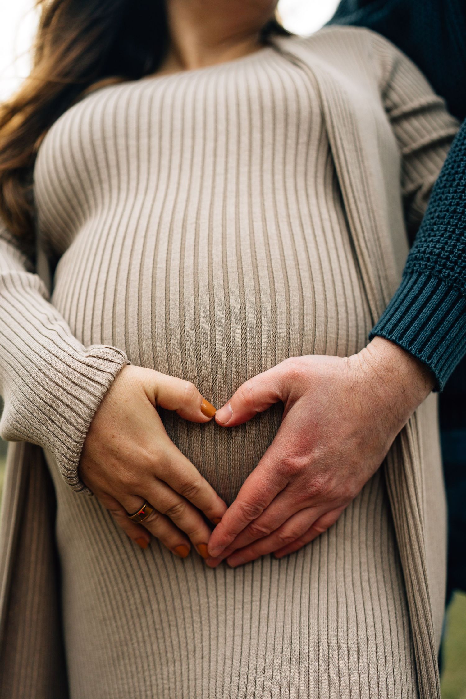 Close up of hands forming heart shape over pregnant belly in cream ribbed sweater.