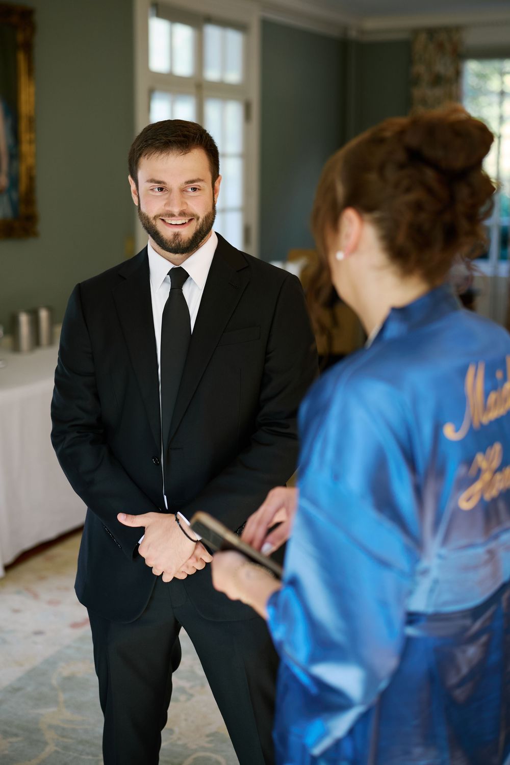 Smiling groom in black suit and tie speaking with bridesmaid in blue satin robe indoors.