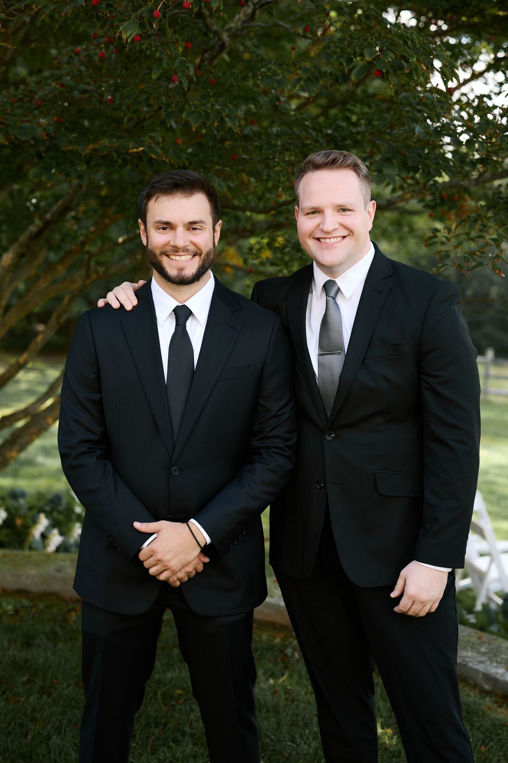 Two groomsmen in black suits smiling outdoors at a wedding ceremony venue surrounded by lush greenery.