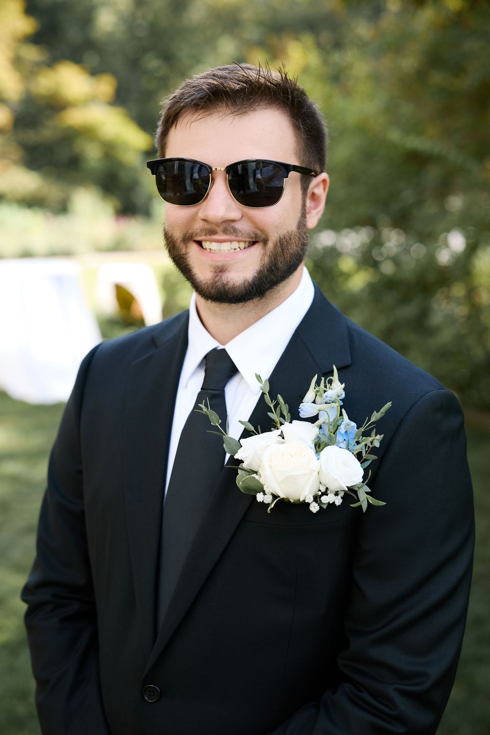 Handsome groom in black suit with floral boutonniere wearing sunglasses outdoors at wedding.