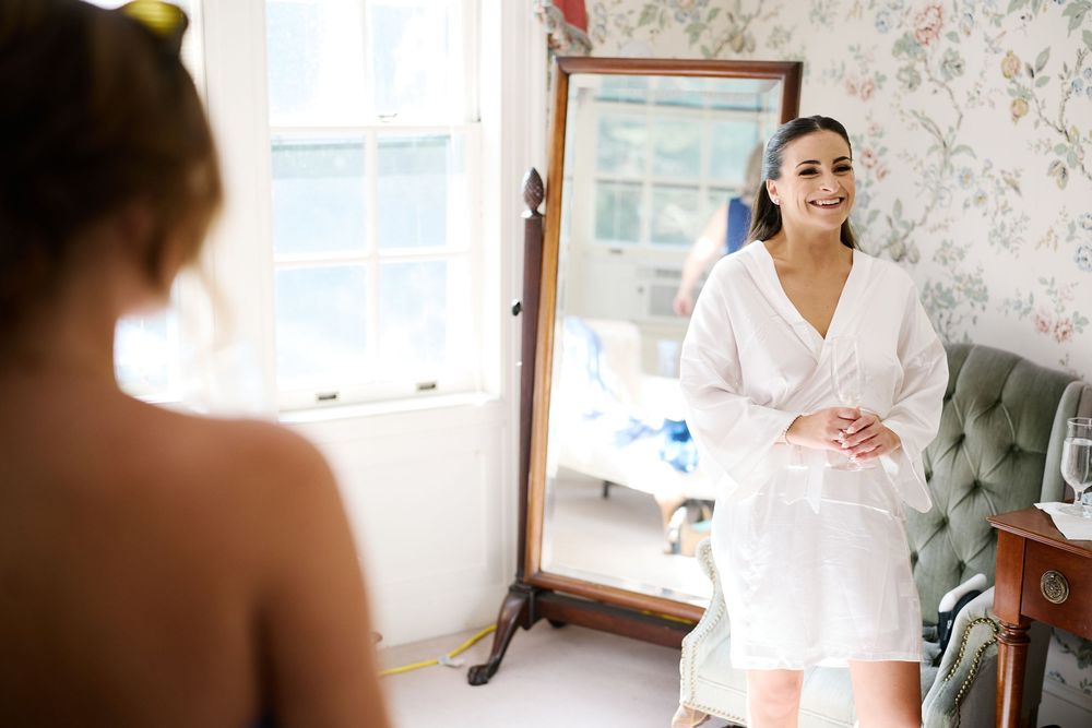 Bride in white robe smiling at her reflection in a tall mirror during wedding preparations in a floral room.