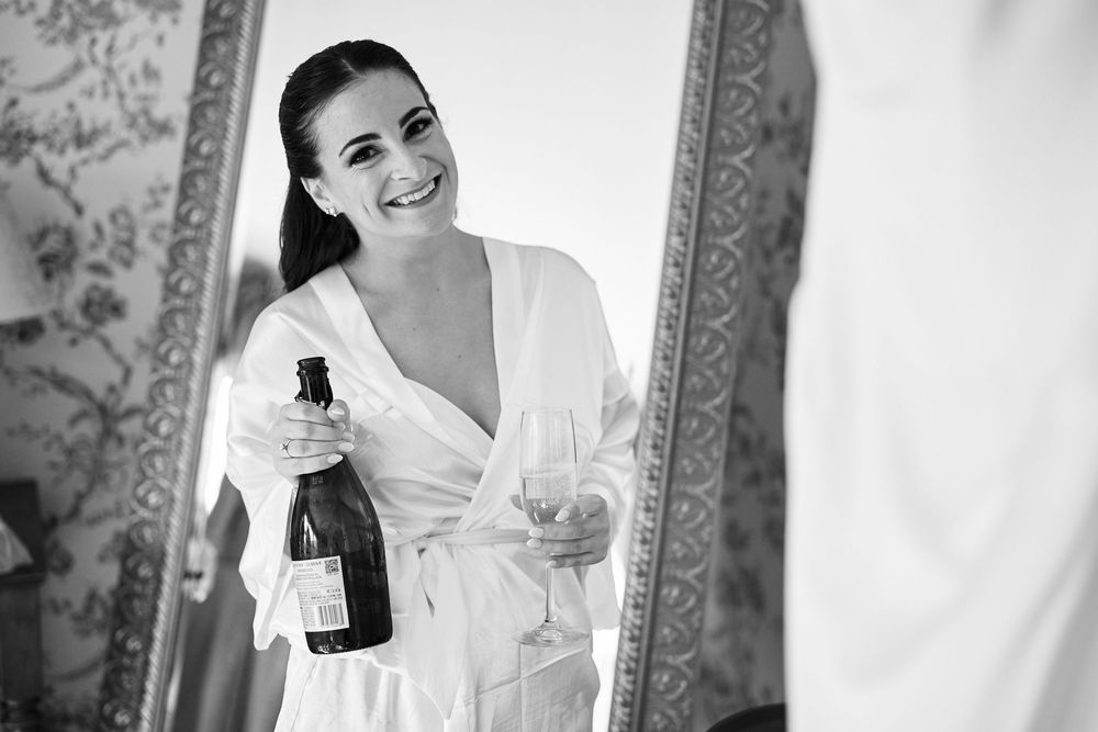 Woman in white robe holding champagne bottle and glass, smiling at mirror in elegant black and white photo.