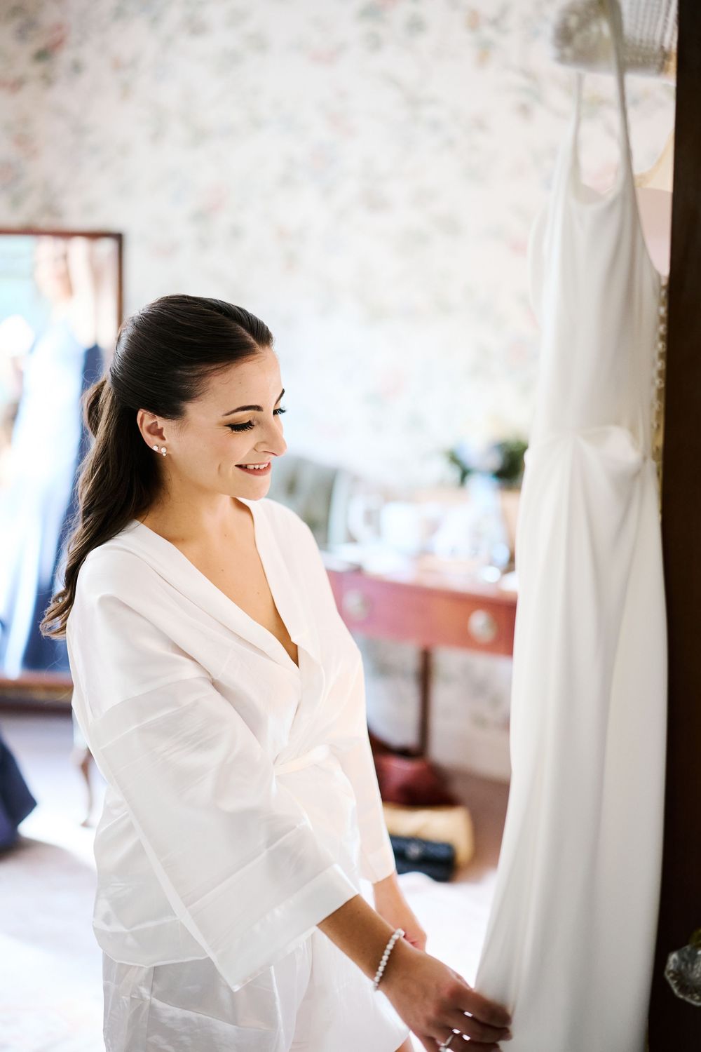 Smiling bride in white robe admiring her wedding dress hanging in a floral wallpapered room.