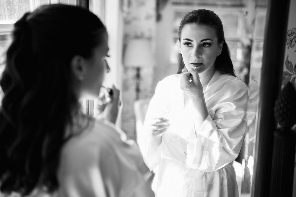 Black and white photo of elegant woman in white robe examining her makeup in an ornate mirror at theBradley Estate in Canton, Massachusetts.