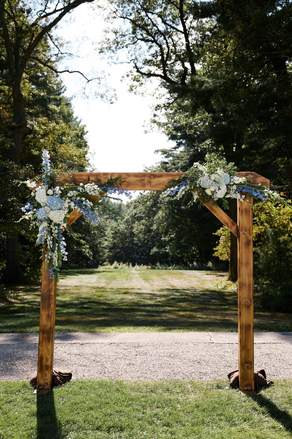 Rustic wooden wedding arch adorned with blue and white flowers, set on a gravel path surrounded by lush green trees.