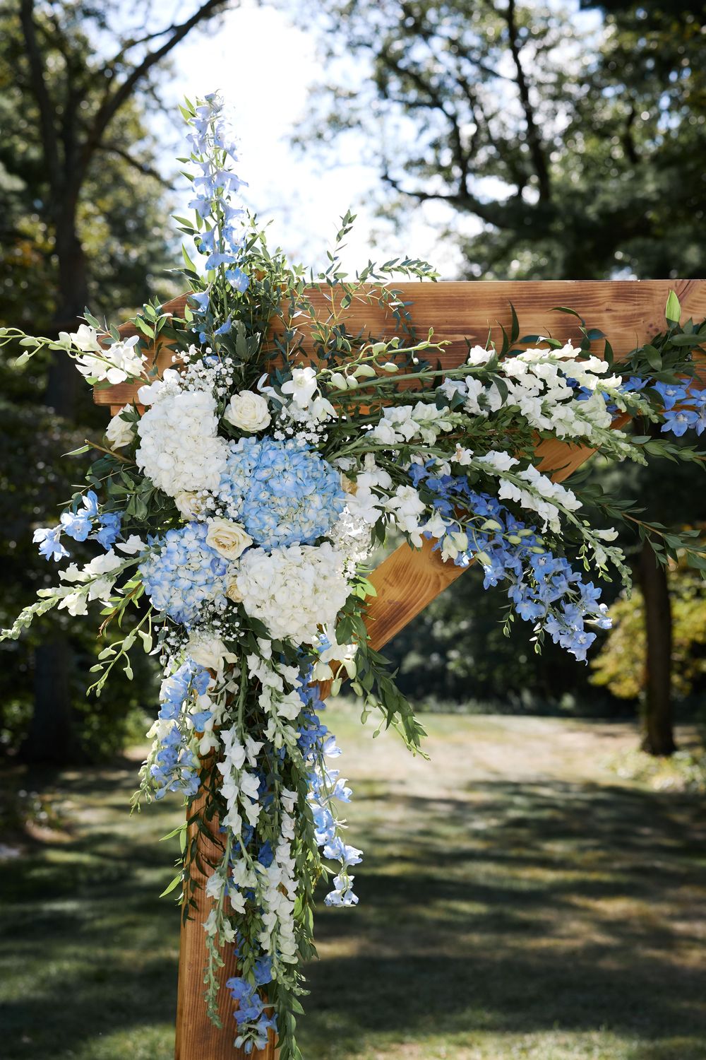 Wooden wedding arch adorned with blue hydrangeas, white roses, and cascading greenery in an outdoor garden setting.