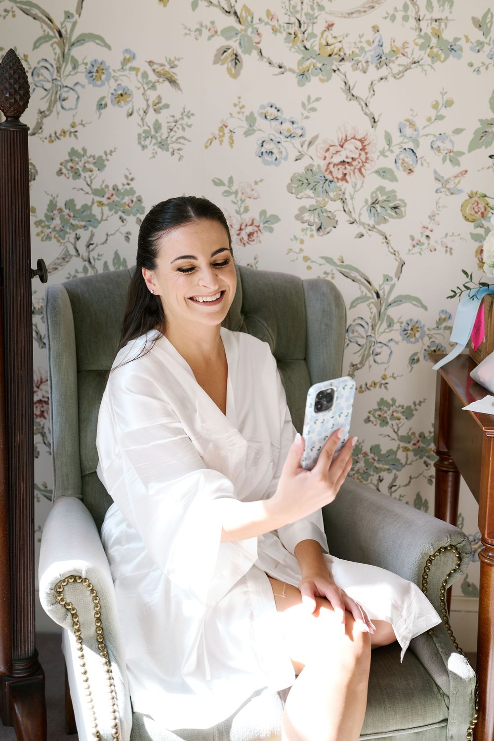 Smiling bride in white robe sitting in armchair looking at phone in room with floral wallpaper.