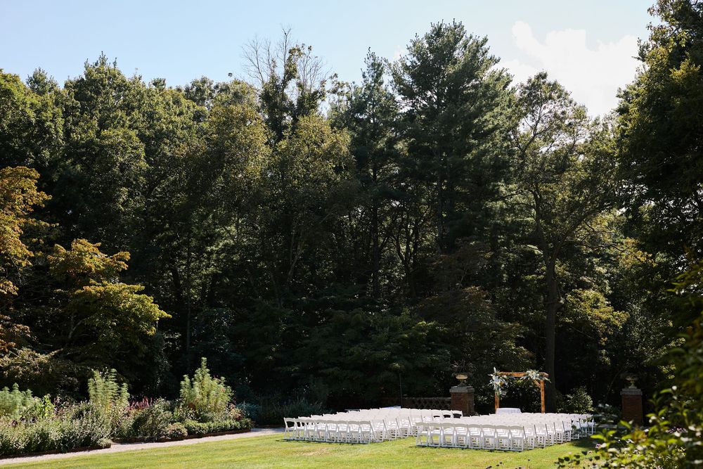 Outdoor wedding ceremony setup with white chairs on green lawn surrounded by lush tall trees on a sunny day at the Bradley Estate.
