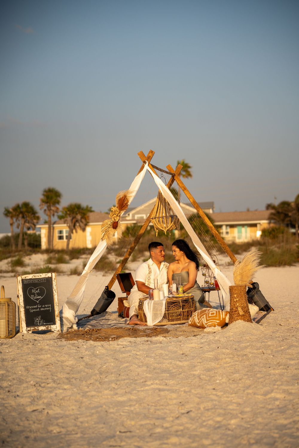 Couple sharing a romantic beach picnic under a boho teepee setup at golden hour on white sand.