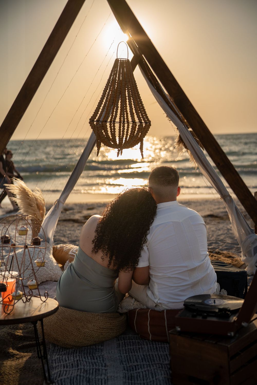 Couple sitting together under a boho triangular arch on the beach at sunset, enjoying a romantic seaside picnic.