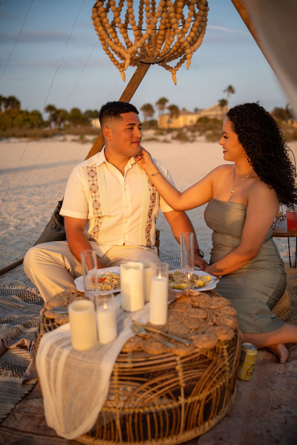Couple sharing a romantic beach picnic at sunset with candles and food on a wicker table.