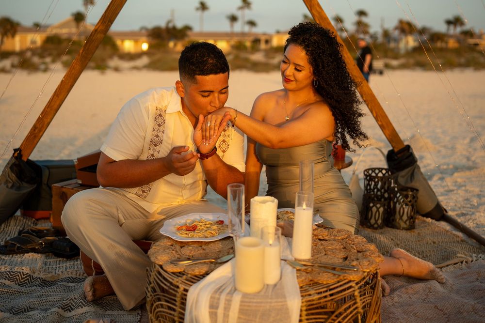 Couple enjoying a romantic beach picnic at sunset with candles, drinks, and food on a woven table.