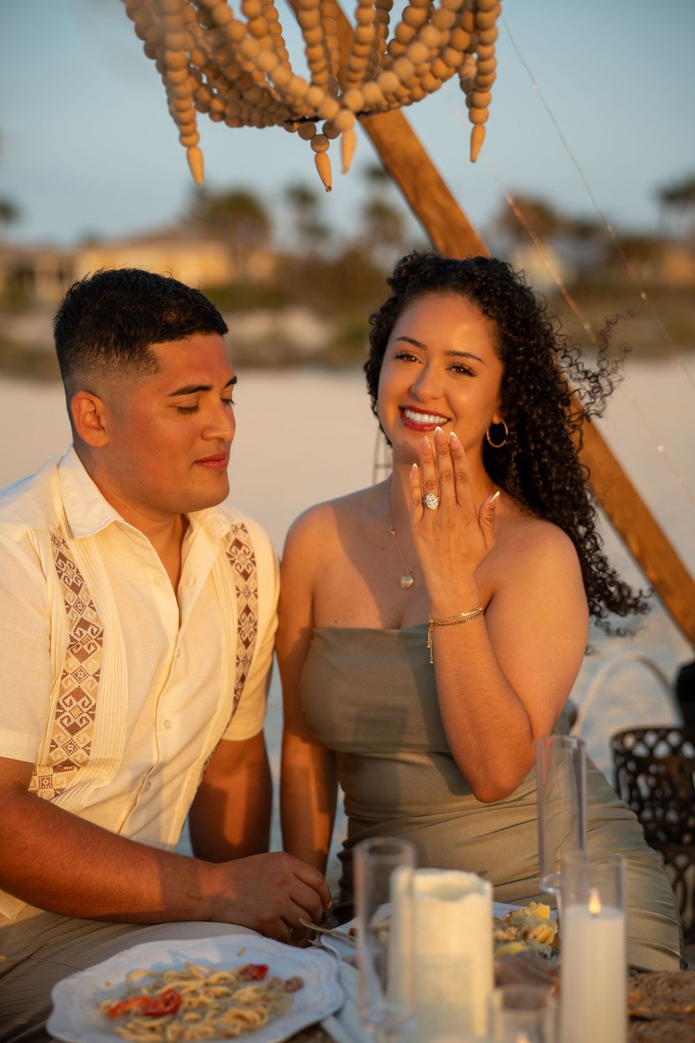 Couple at beach dinner, woman smiling and showing engagement ring, romantic sunset proposal moment.