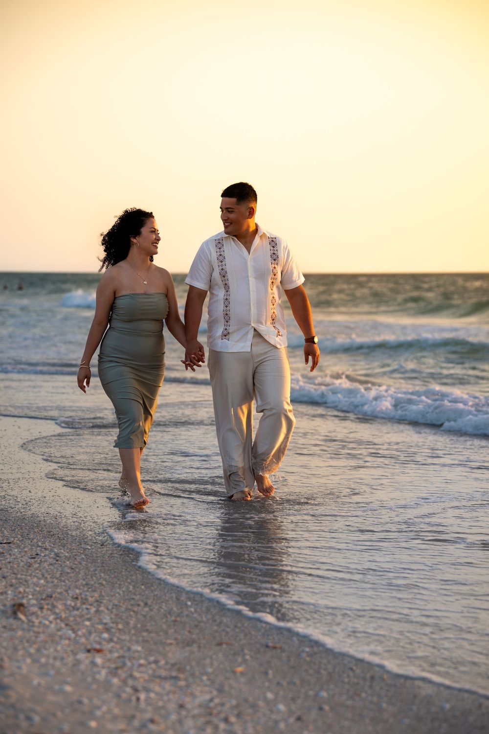 Couple holding hands walking barefoot on a sandy beach at sunset, waves gently washing over their feet.
