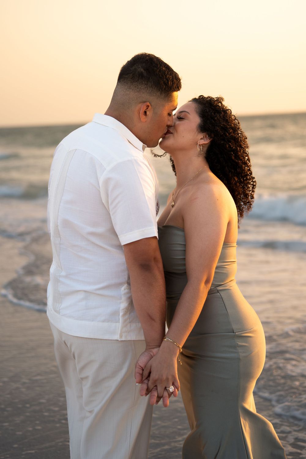 Couple sharing a romantic kiss on the beach at sunset, holding hands, woman in olive dress, man in white shirt.