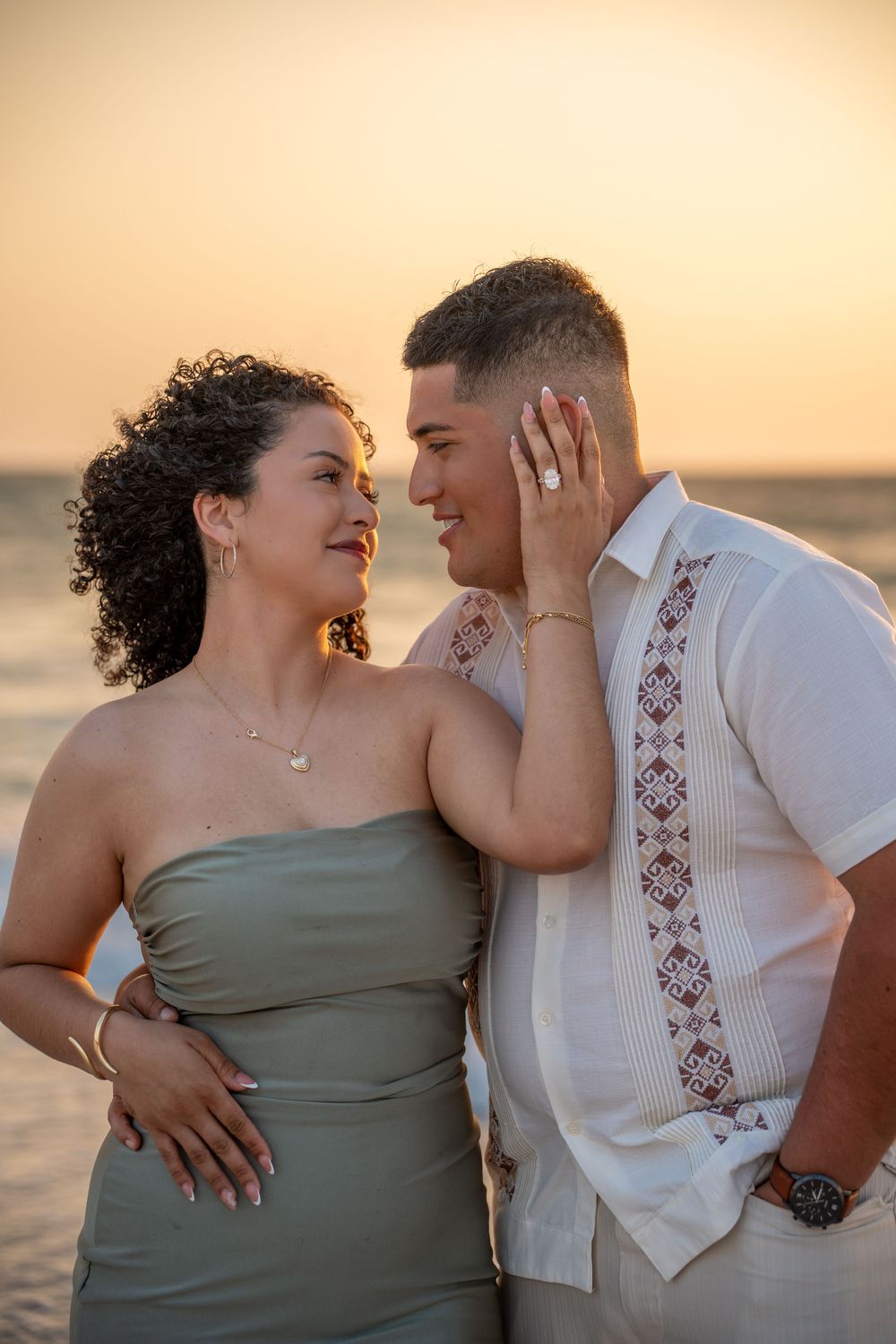 Couple sharing a romantic sunset beach moment, woman in sage strapless dress showing engagement ring, man in embroidered guayabera shirt.
