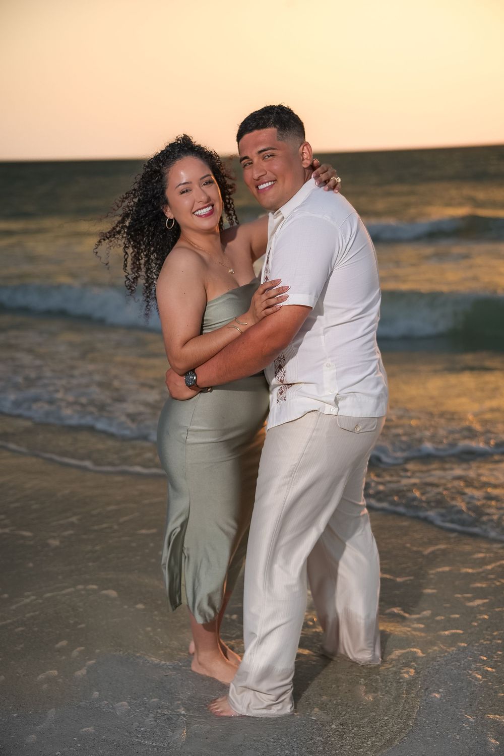 A smiling couple embraces on a sandy beach at sunset, woman in green dress, man in white shirt and tan pants.