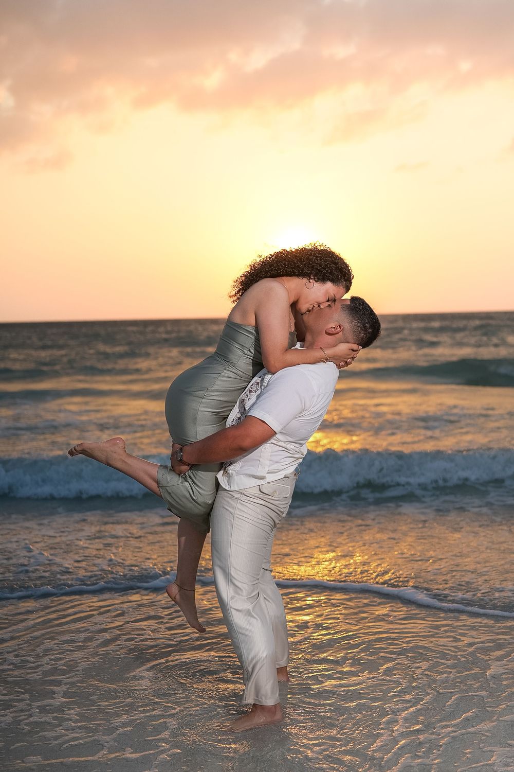 Romantic couple embracing on a beach at sunset, man lifting woman in his arms with ocean waves behind them.
