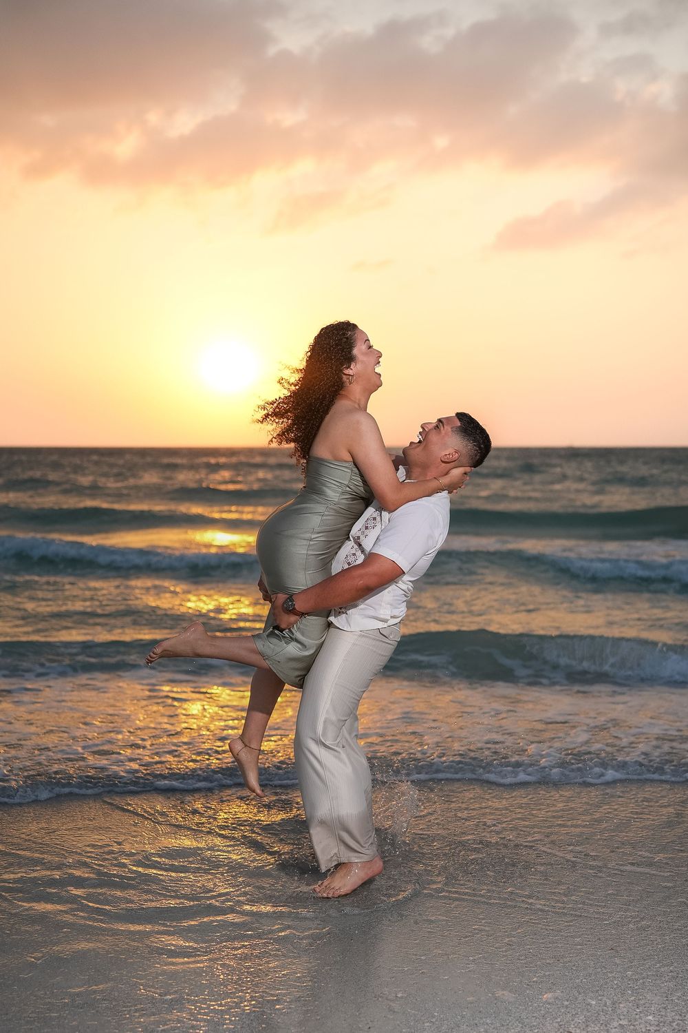 Couple embracing romantically on a beach at sunset, man lifting woman in a silver dress with ocean waves behind them.
