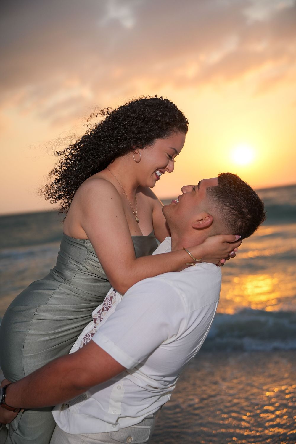 Joyful couple embracing at golden sunset beach, woman laughing while being lifted by man in white shirt.