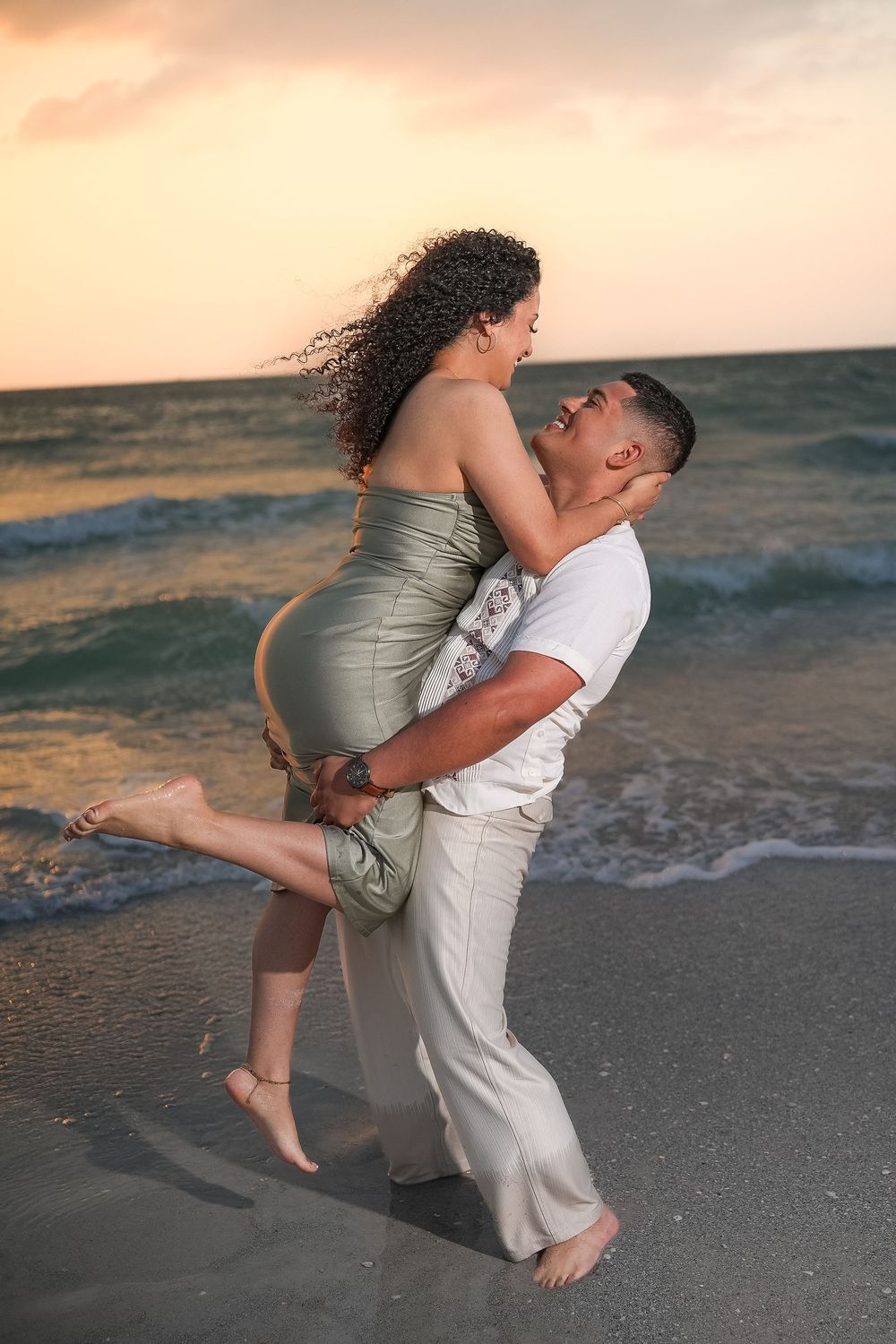 A couple shares a romantic moment on the beach at sunset, man lifting woman in green dress by ocean waves.