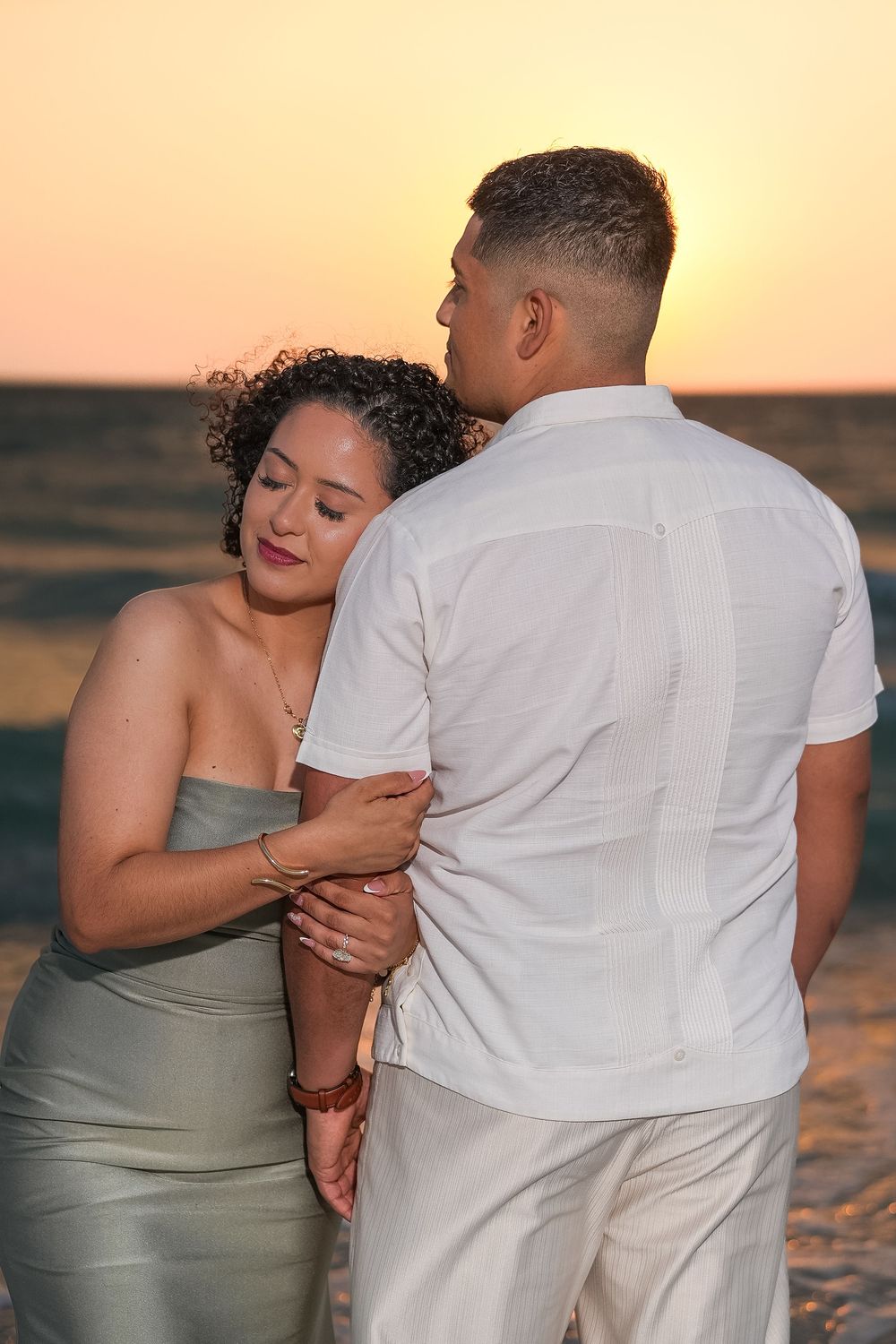 Couple embracing at sunset on the beach, woman in gray dress, man in white shirt, golden sky over ocean.