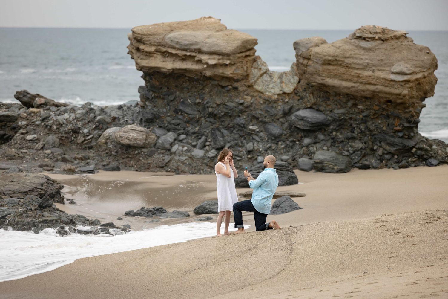 Romantic beach proposal with man on one knee before woman, dramatic rock formations behind them.