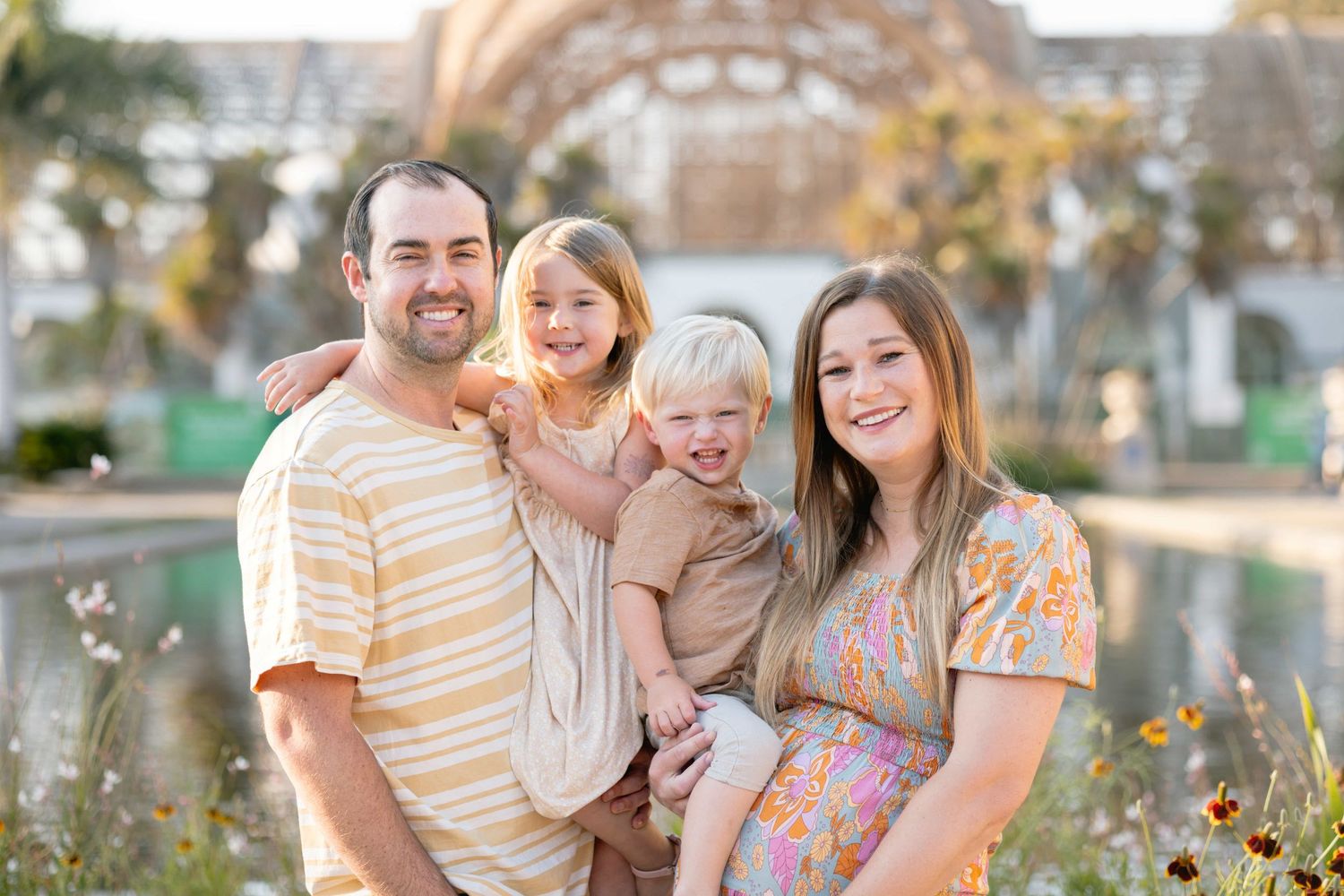 Happy family of four poses outdoors, pregnant mother in floral dress with father and two young children smiling.