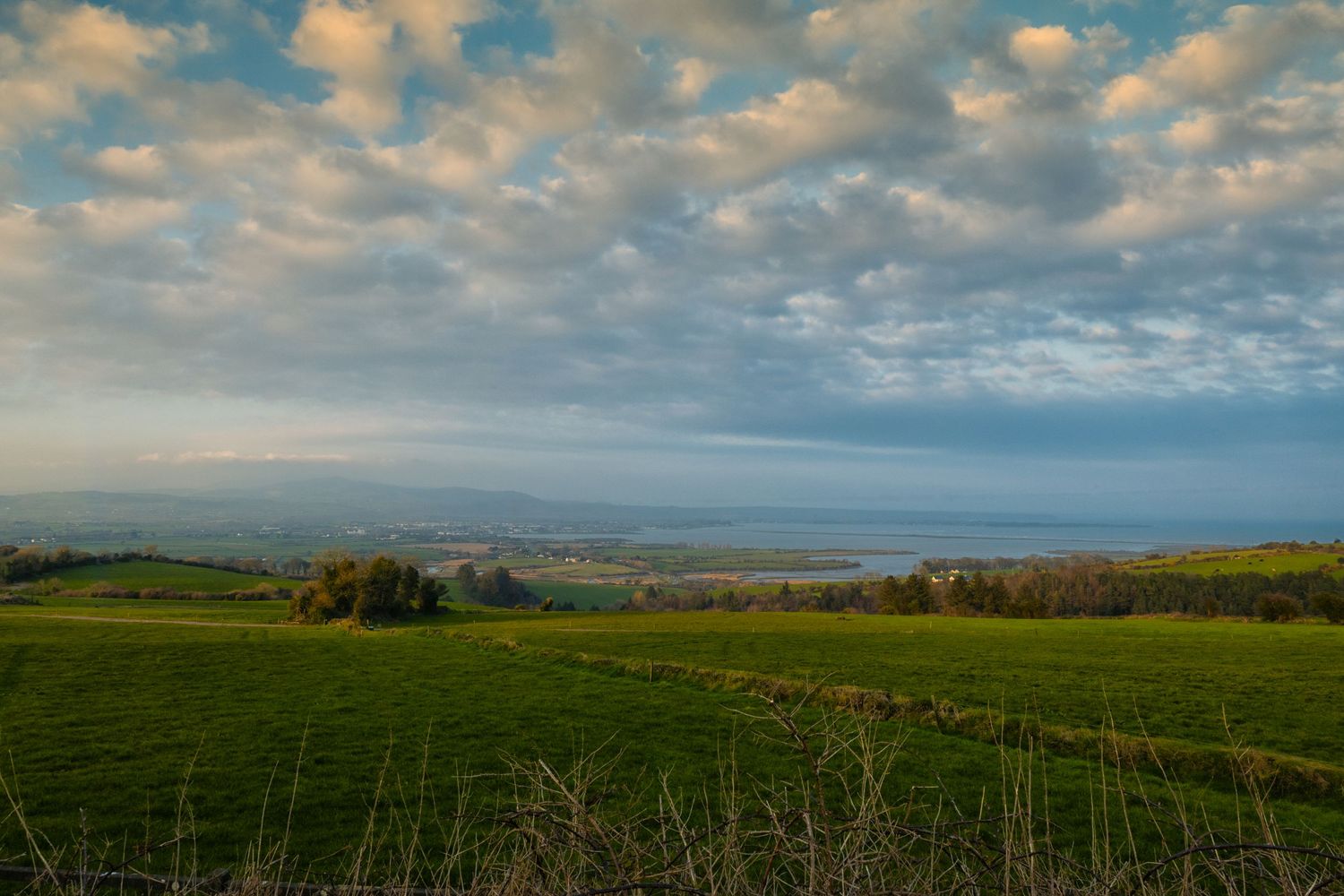 Lush green fields stretch toward a distant coastline under a dramatic sky with scattered clouds at golden hour.