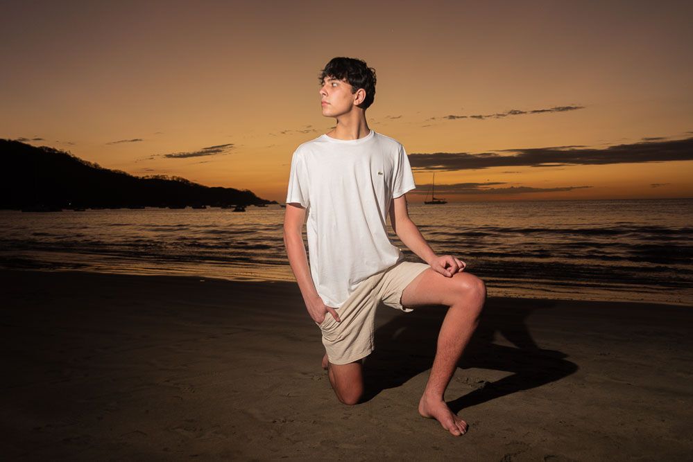 Young man in white t-shirt and beige shorts poses on sandy beach at golden sunset with ocean backdrop.