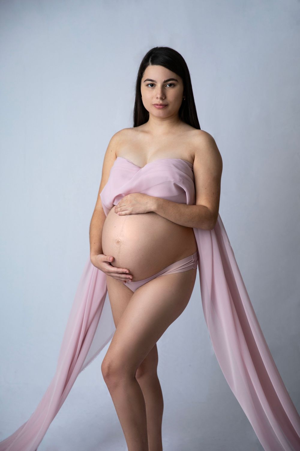 Pregnant woman in pink fabric drape holding baby bump in studio maternity photo shoot on light gray background.