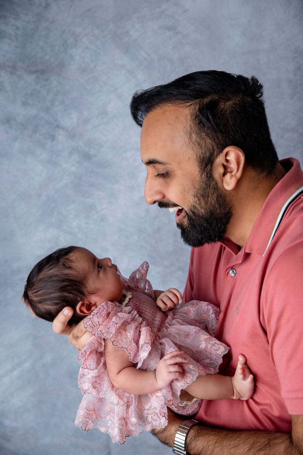 A bearded man in a pink shirt smiling at a newborn baby girl in a pink lace dress cradled in his arms.
