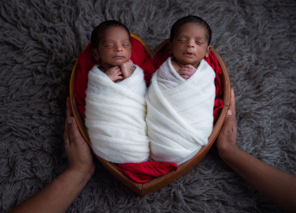 Two newborn twins swaddled in white wraps sleeping in a heart-shaped wooden basket on a gray fur background.
